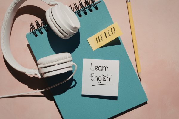 From Basic to Advanced: How to Master English Grammar in the U.S. 7 Notebook with Learn English note, headphones, pencil, and sticky note saying Hello on a pink background.