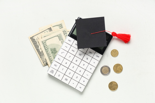 The 2026 U.S. Tuition Fee Report: Trends in Public vs. Private University Costs 4 A top-down view of a white calculator with a small black graduation cap on top, surrounded by U.S. dollar bills and coins, representing the calculation of university costs.