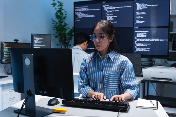 Cybersecurity Management: Why a Master's Degree is the Key to Senior Executive Roles 10 Woman programming at a computer in an office with large screens displaying code in the background.