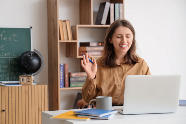 How to Improve Your English Vocabulary Quickly: Intensive ESL for Beginners 8 Smiling woman waving during a video call at home office, laptop, books, and a chalkboard in the background.