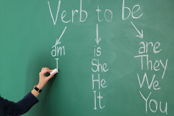 How to Improve Your English Vocabulary Quickly: Intensive ESL for Beginners 9 Person writing verb conjugation to be on chalkboard with am, is, are examples.