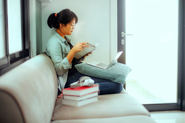 How to Improve Your English Vocabulary Quickly: Intensive ESL for Beginners 7 Young woman studying on a sofa with books and a laptop by a bright window.