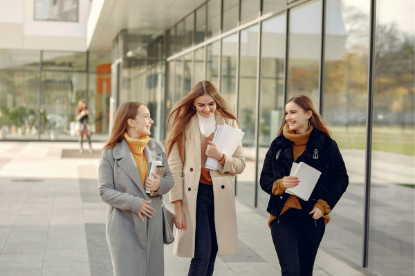 Balancing Work and Study: A Commuter Student’s Guide 8 Three young women walking and talking outside a modern glass building, holding notebooks and smiling.