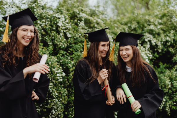 Happy graduates in caps and gowns celebrate with diplomas in hand, standing outdoors amidst greenery.