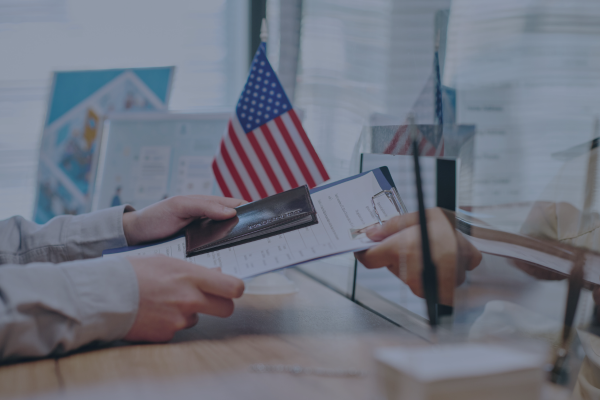 Person hands passport and documents at counter near American flag, representing travel or visa application process.