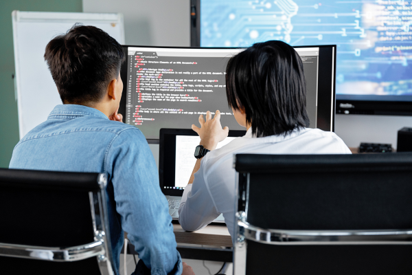 Understanding Cybersecurity Frameworks: Essential Cybersecurity Certification Tips 7 Two developers collaborating on code at a desk with dual monitors displaying programming languages in a modern office.
