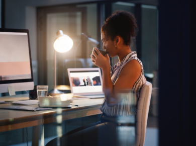 Side Hustles for College Students: Balancing Freelancing with Full-Time Studies 28 Woman working late at an office desk, drinking coffee while using a computer and laptop, with a lamp illuminating her space.