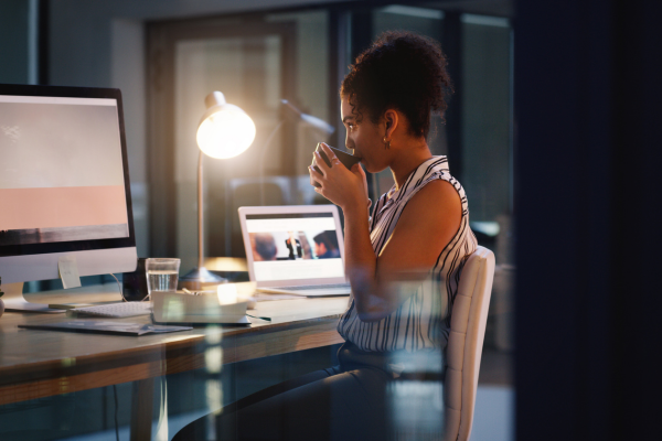 Woman working late at an office desk, drinking coffee while using a computer and laptop, with a lamp illuminating her space.