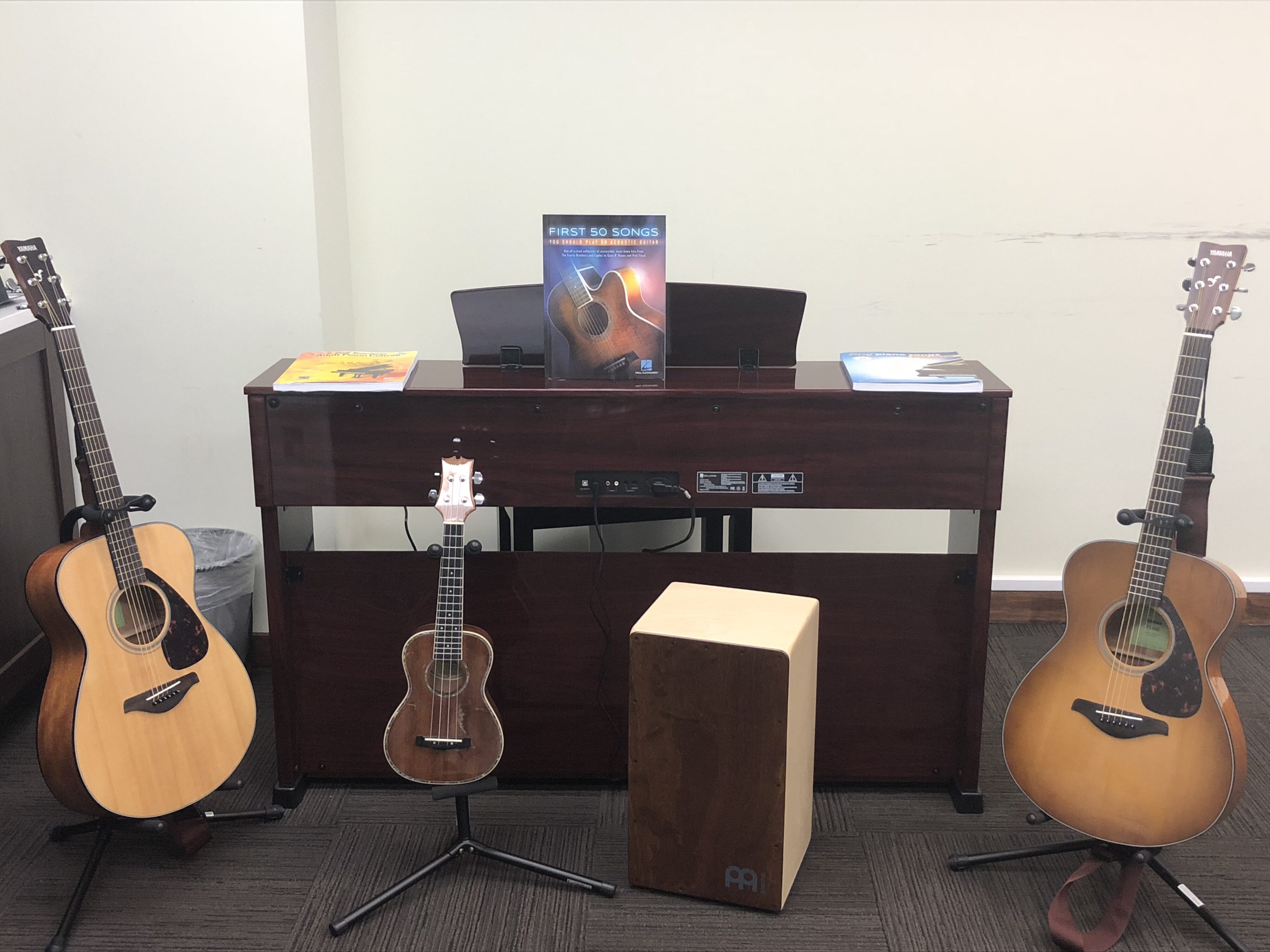 Acoustic guitars, ukulele, and cajon setup in a music room with a keyboard and sheet music.