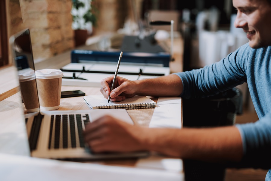 Man writing in notebook at a cafe, laptop and coffee cup on desk, casual work environment.