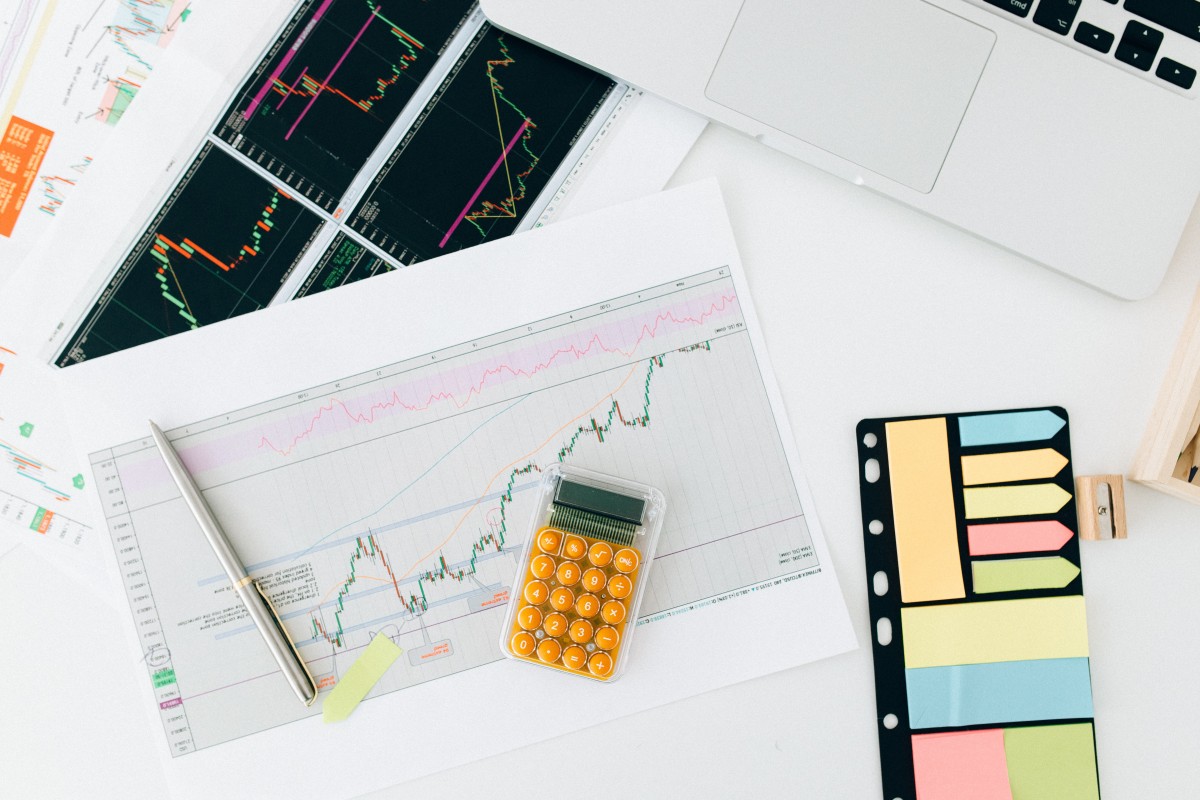 Trading analysis setup with stock charts, a yellow calculator, sticky notes, and a laptop on a white desk.