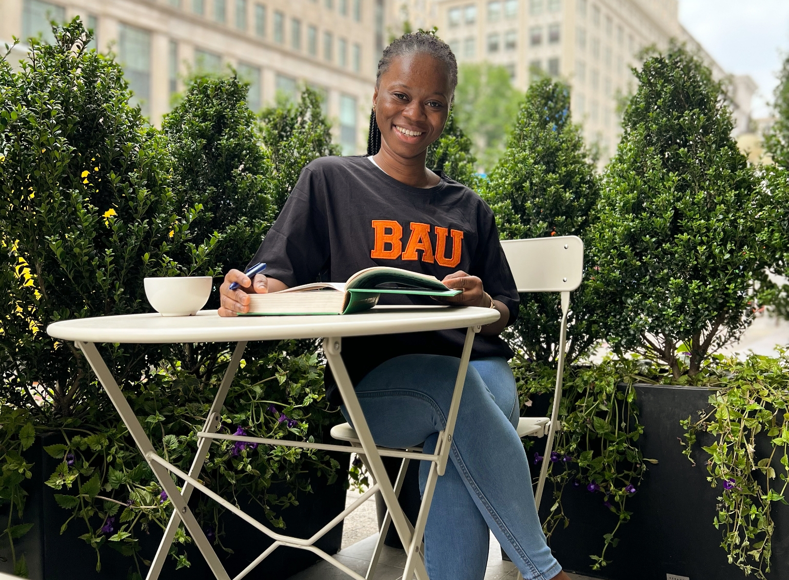 Smiling woman studying at an outdoor cafe table, surrounded by greenery, with a book and a cup.