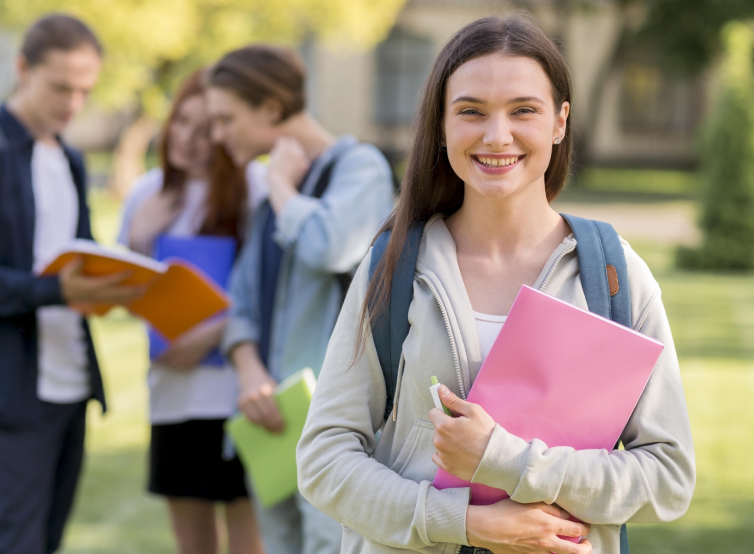Smiling student holding books in a campus setting with peers discussing in the background.
