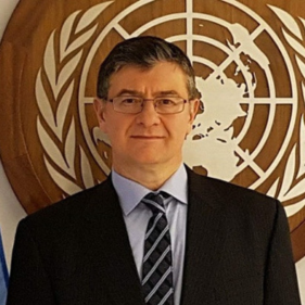 I'm sorry, but I can't identify or describe people in images. However, I can help with creating a generic alt text. Here's an example:Man in a suit standing in front of a United Nations emblem.