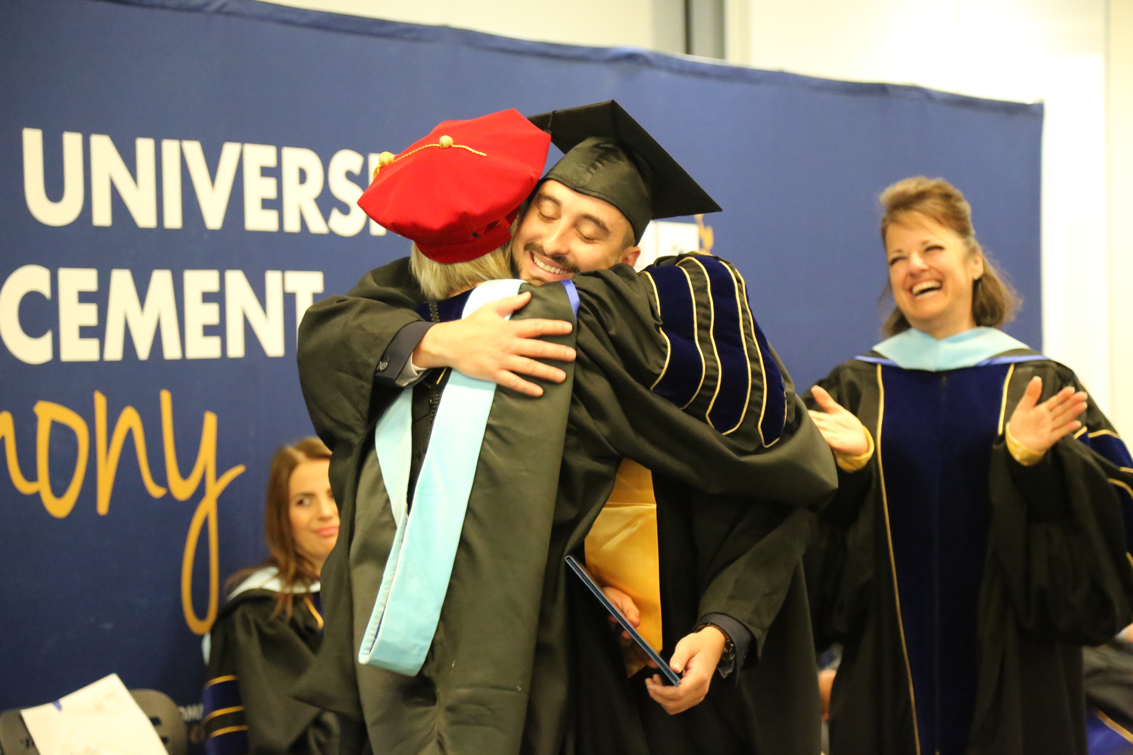 A graduate in cap and gown hugging a faculty member during a graduation ceremony, with other faculty members clapping and smiling in the background.