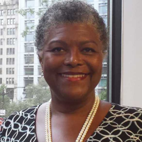 Smiling person in an office setting with city buildings in the background, wearing a patterned outfit and a pearl necklace.