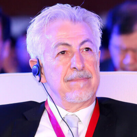 Elderly man in a suit with a headset and red lanyard, attending an event, looking thoughtfully upward.