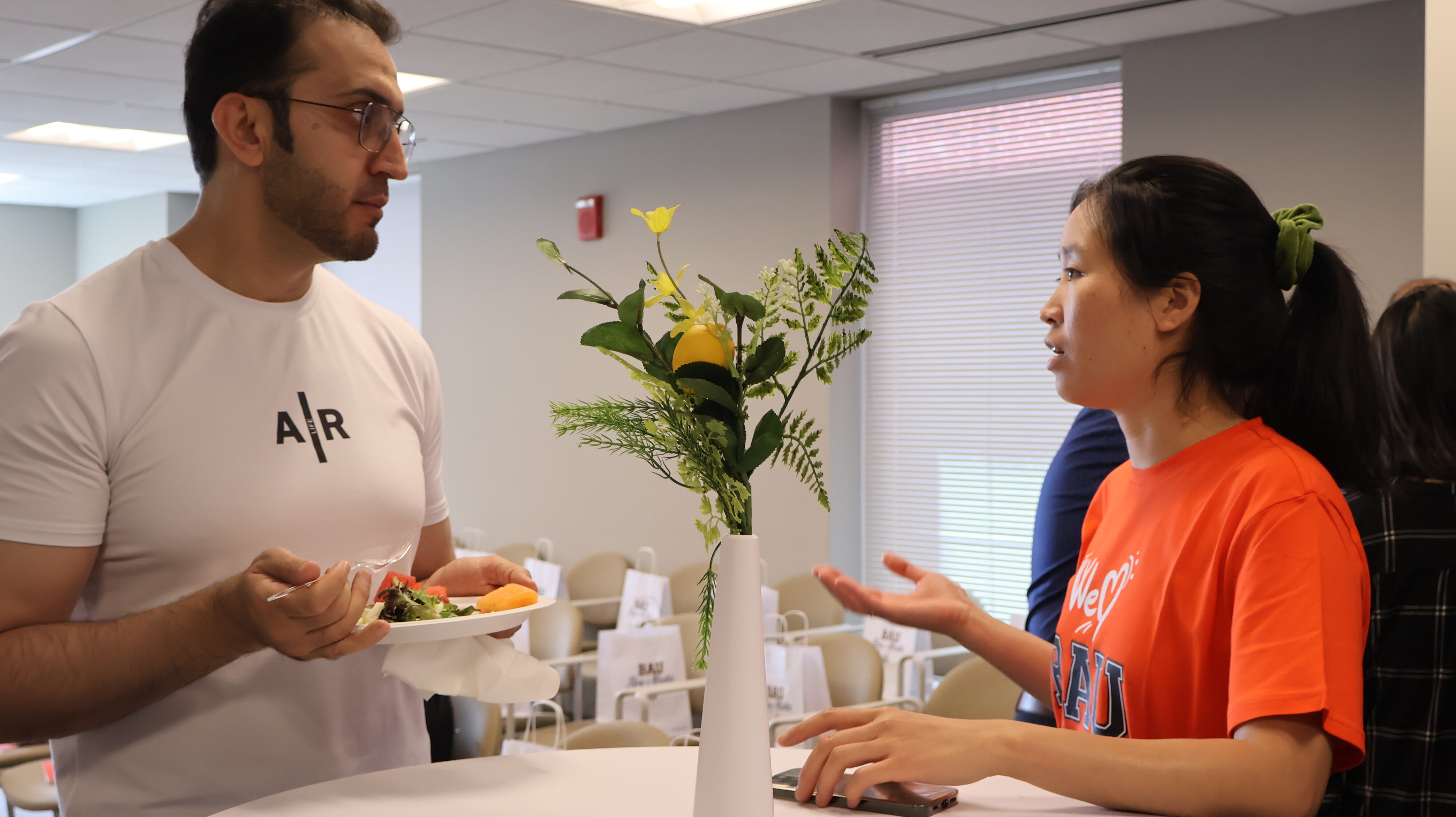 Two people having a conversation at a table with a vase, one holding a plate of food.