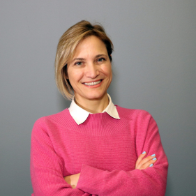 Smiling person in a pink sweater standing against a gray background with arms crossed.