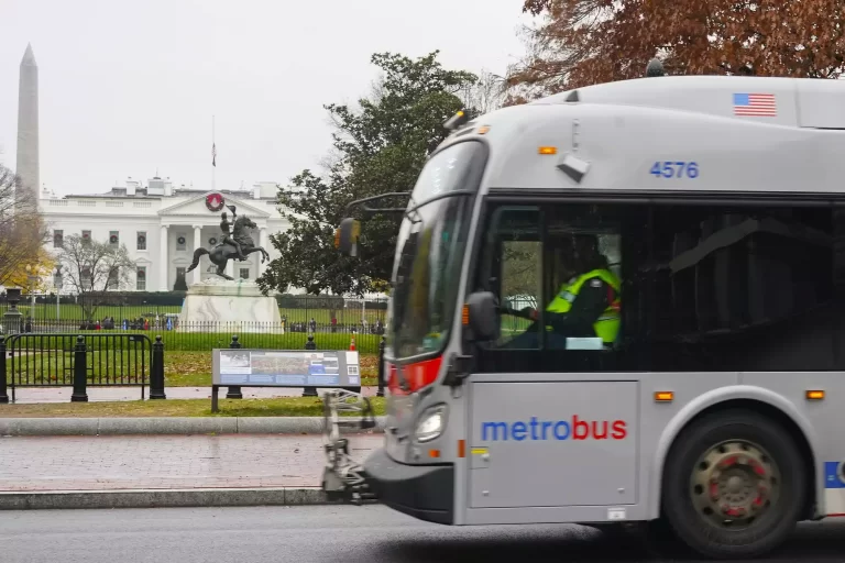 Metrobus passes by the White House with Washington Monument in the background on a cloudy day.