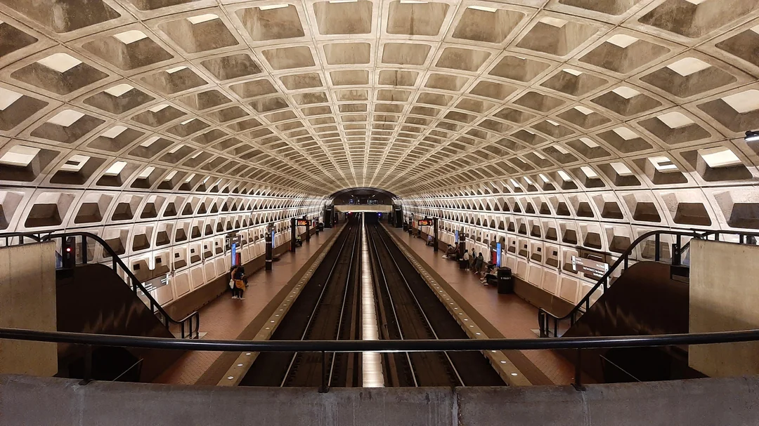 Underground metro station with a curved concrete ceiling, empty tracks, and waiting passengers on the platform.