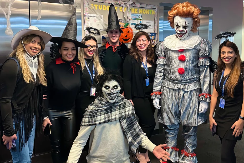 Office Halloween party with creative costumes, featuring witches, angels, and a spooky clown, posing for a group photo.