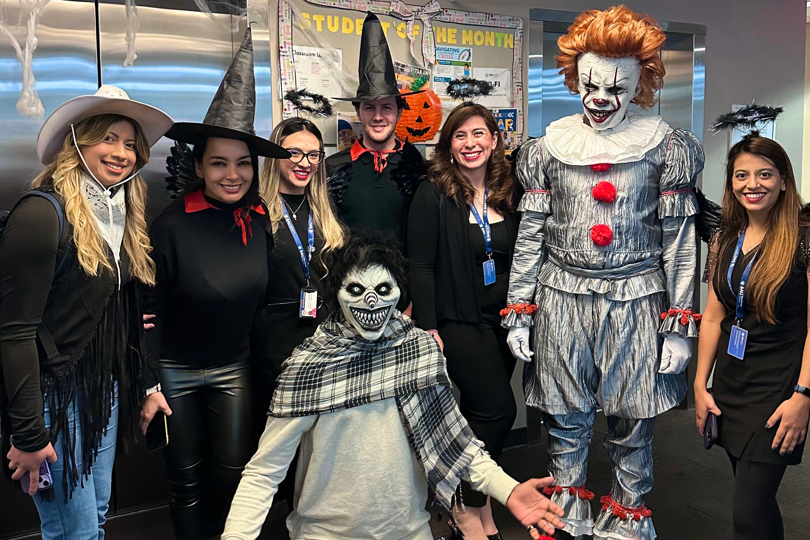 Office Halloween party with creative costumes, featuring witches, angels, and a spooky clown, posing for a group photo.