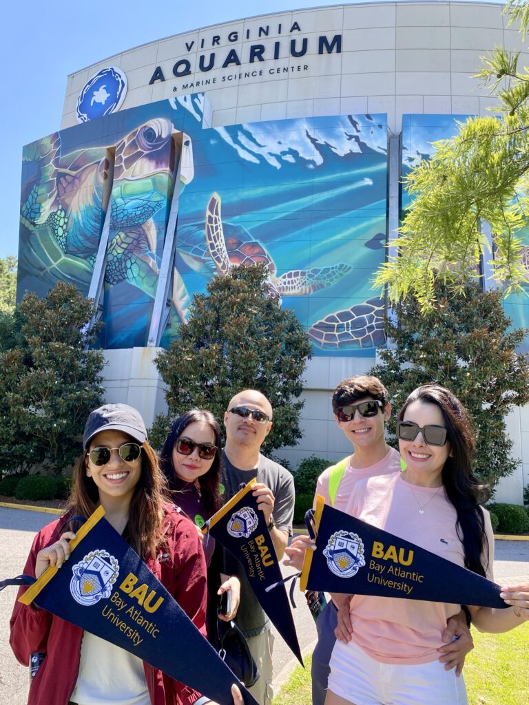 20240625_185039312_iOS Group at Virginia Aquarium with BAU flags, smiling in front of vibrant sea turtle mural on a sunny day.