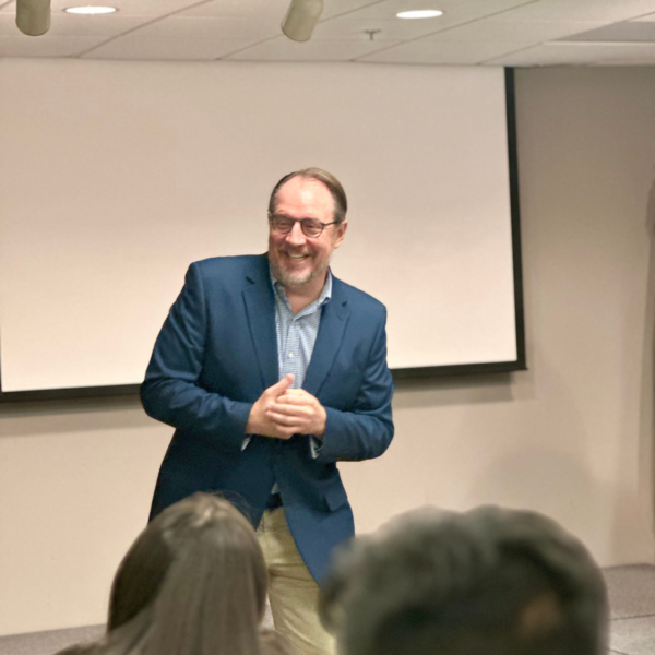 A professor in a blue blazer smiling while addressing an audience during an indoor presentation.