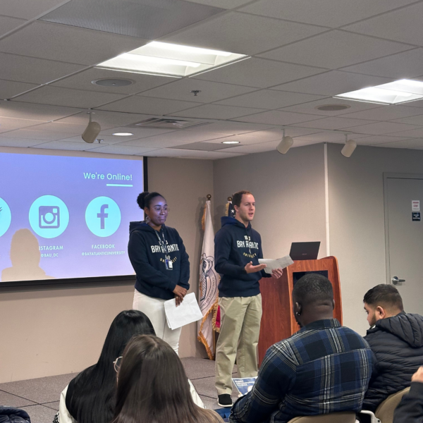 Two presenters wearing BAU hoodies speaking in front of an audience during an indoor presentation.