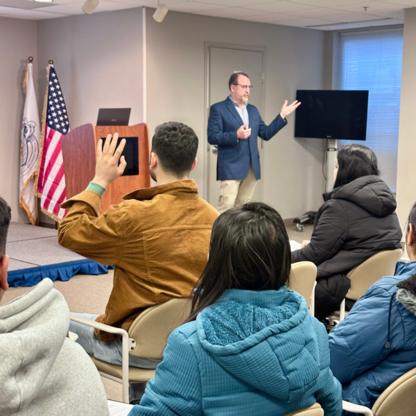 A speaker presenting to an audience in a room, with one audience member raising their hand. An American flag and a television screen are visible in the background.