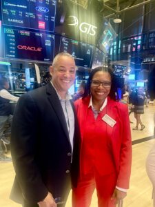 A man and woman wearing suits pose in front of several large screens displaying share prices at the New York Stock Exchange.