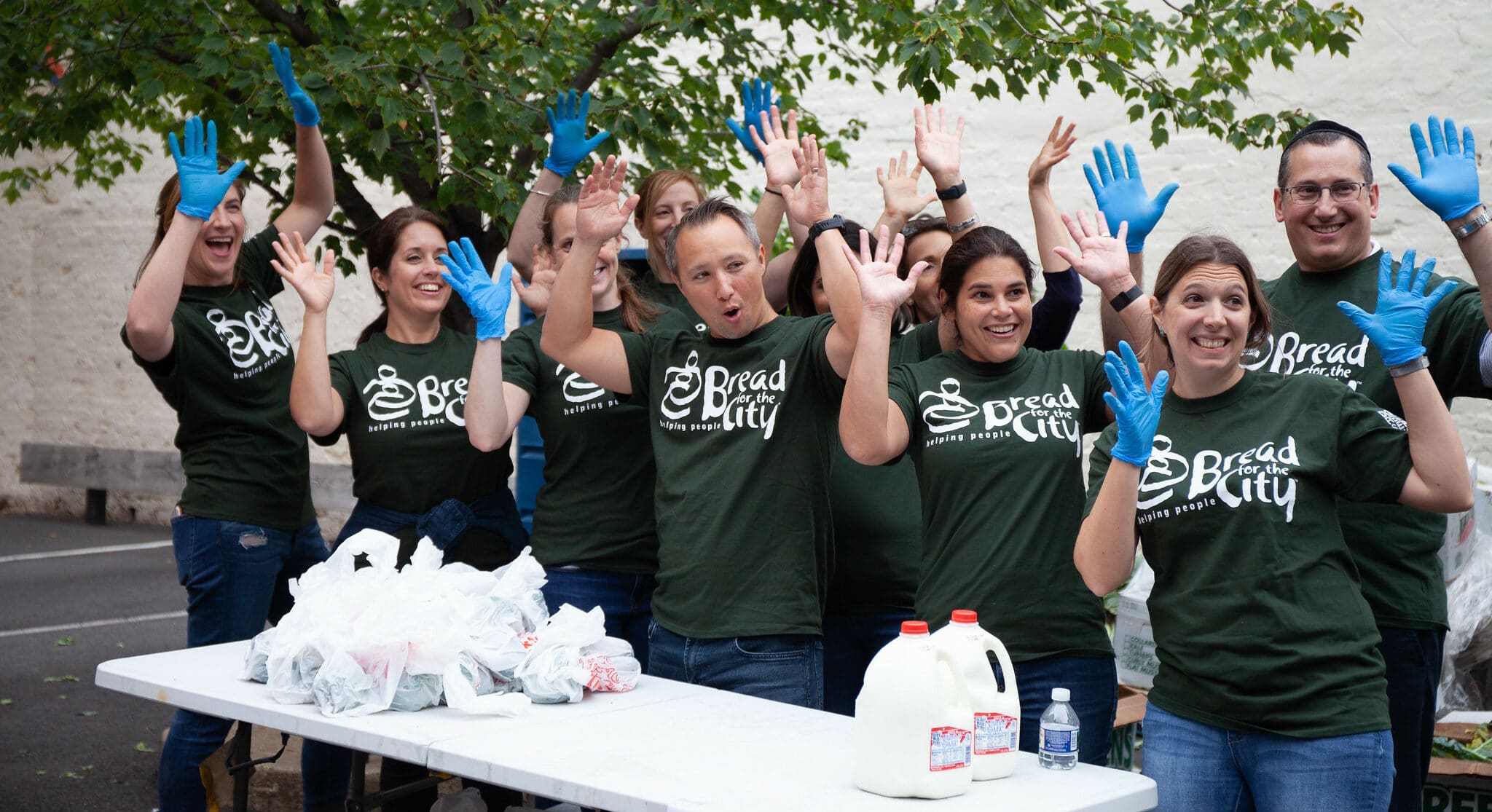 Volunteers in green shirts and gloves cheer at a community service event with food donations on a table.