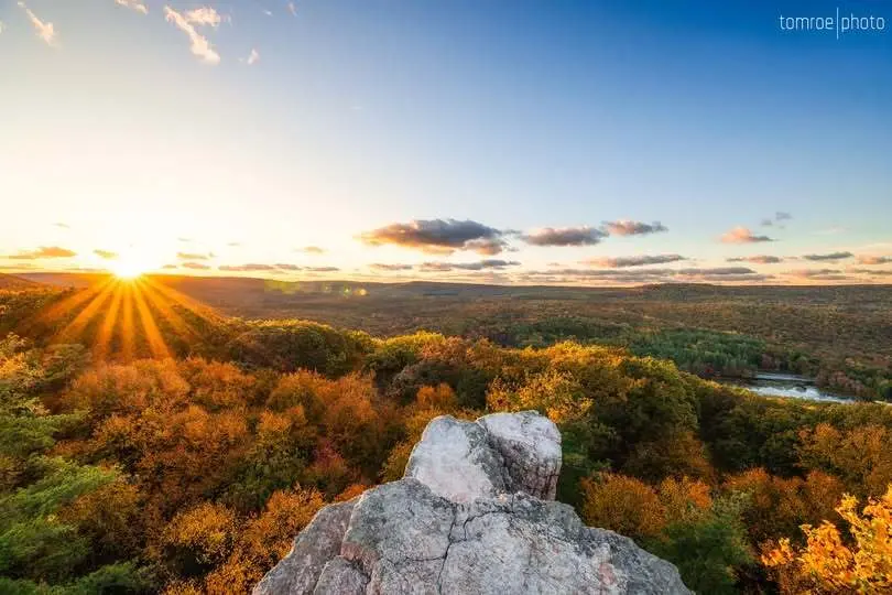 Scenic view of a sunrise over autumn forest from a rocky cliff, with a clear blue sky and sun rays peeking through clouds.