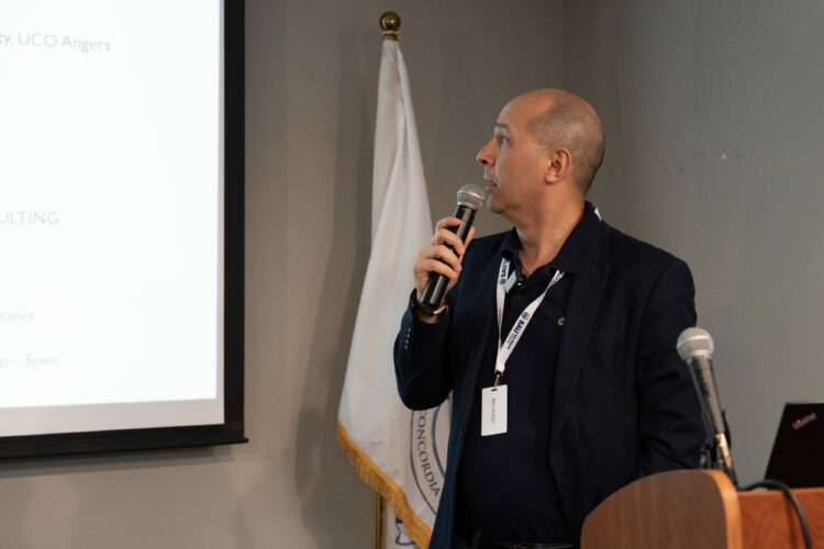 Speaker presenting at a conference, using a microphone and pointing to a slide on a screen.