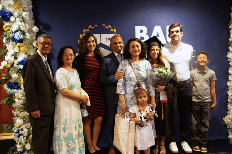 Family celebrating graduation ceremony with a graduate in cap and gown, holding flowers, smiling at the camera.