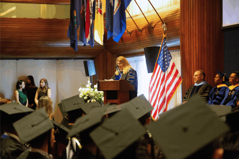 Graduation ceremony with speaker at podium, graduates in caps, and flags in background.