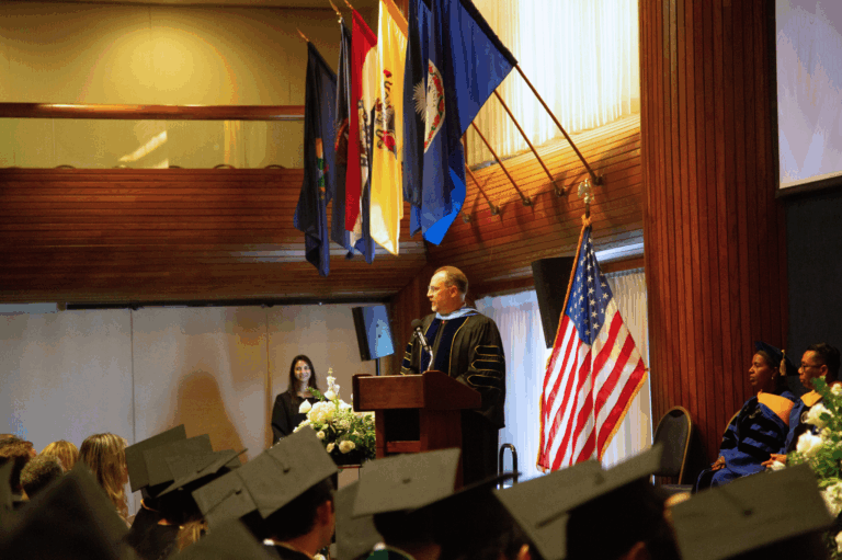 Speaker addressing graduates at a ceremony, with national flags and an audience in caps and gowns.