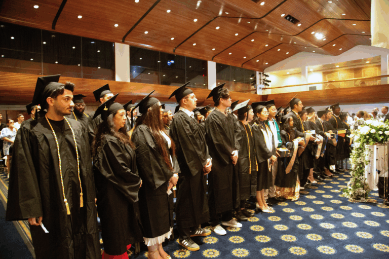 Graduates in caps and gowns standing at a ceremony in a large hall with a floral arrangement.
