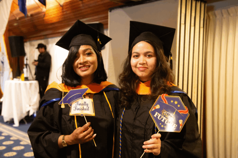 Two graduates in caps and gowns celebrating with Finally Finished and Bright Future signs at their commencement ceremony.