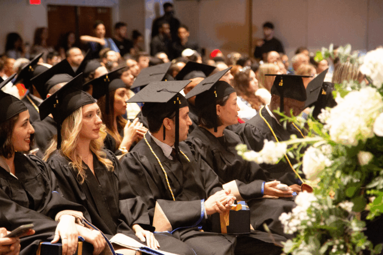 Graduates in caps and gowns seated at a commencement ceremony, holding diplomas and smiling eagerly.