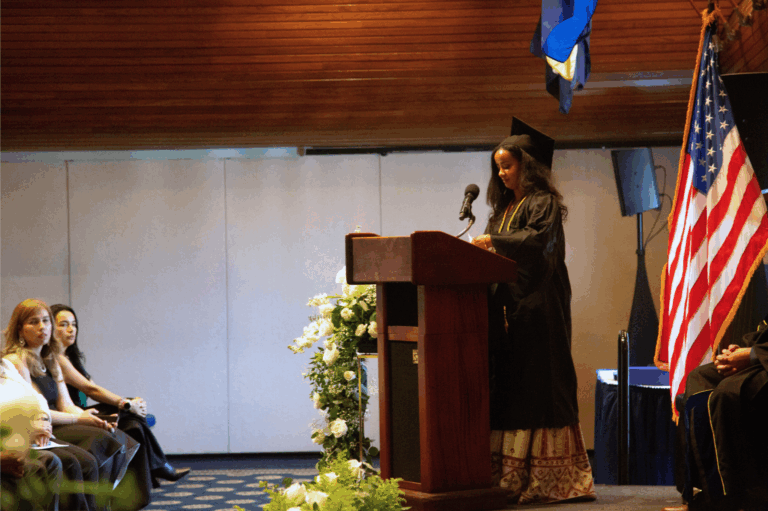 Graduation speaker in cap and gown delivers speech at podium with audience and American flag in background.
