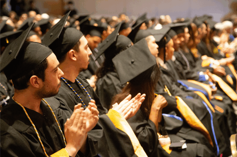 Students in caps and gowns at a graduation ceremony, clapping during the event.