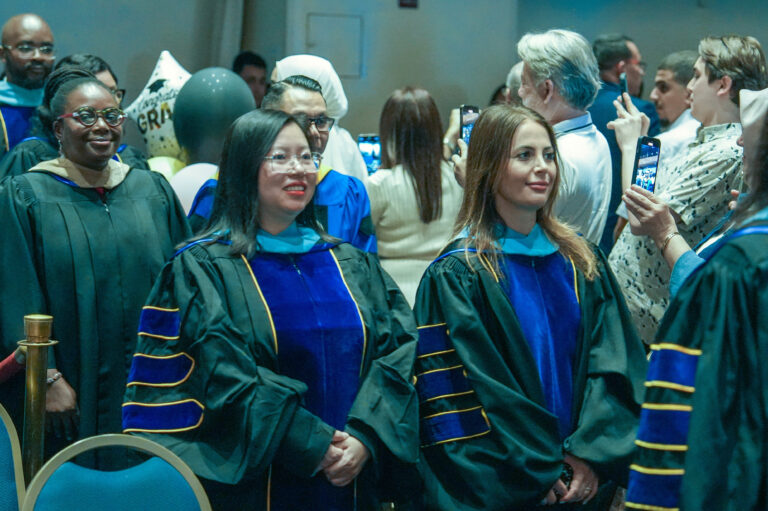 Graduates in academic regalia stand proudly at a commencement ceremony, surrounded by friends taking photos.