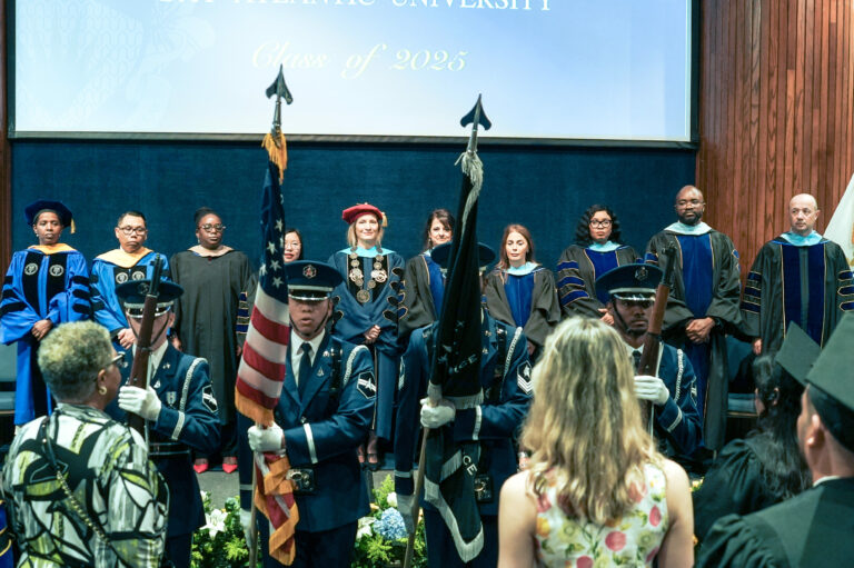 Graduation ceremony with university faculty in robes and honor guard presenting flags, Class of 2025 banner above.