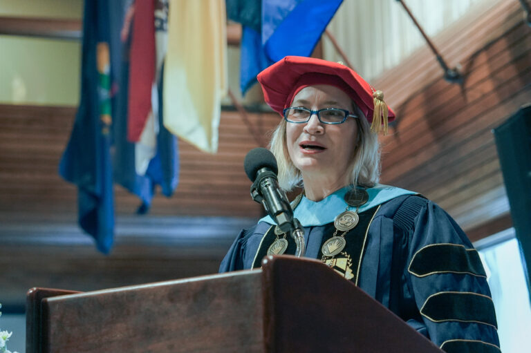 Speaker in academic regalia with chains at podium, delivering speech at a graduation ceremony.