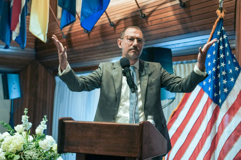 Man giving a speech at a podium with flags in the background, making gestures with arms raised.