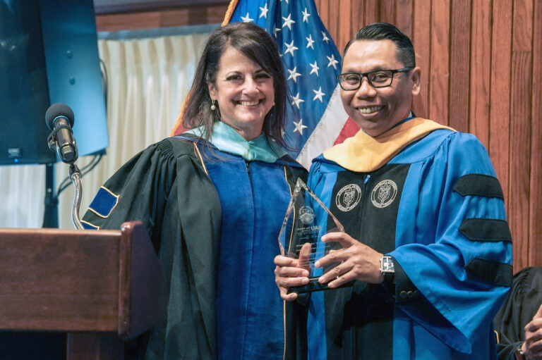Two people in academic gowns smiling and holding an award at a ceremony with an American flag in the background.