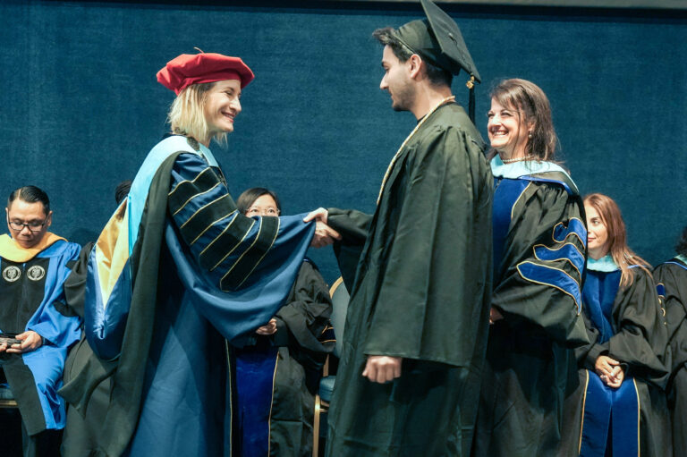 Graduate shaking hands with professor at ceremony, wearing caps and gowns, with faculty seated in the background.