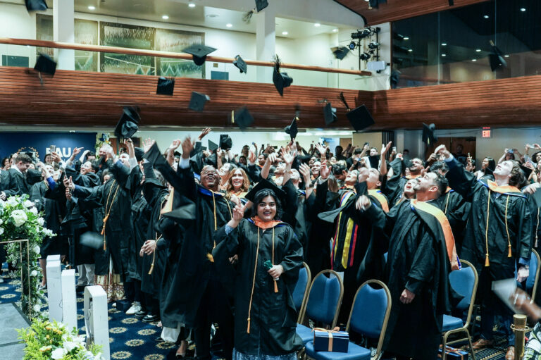 Graduates celebrate by tossing caps in the air at a commencement ceremony in an indoor auditorium.